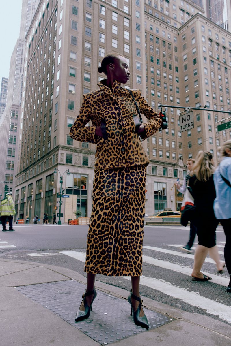 Fashion shot in urban setting shows a black model in an animal print skirt suit on a city street with surrounding buildings.
