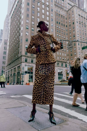 Fashion shot in urban setting shows a black model in an animal print skirt suit on a city street with surrounding buildings.