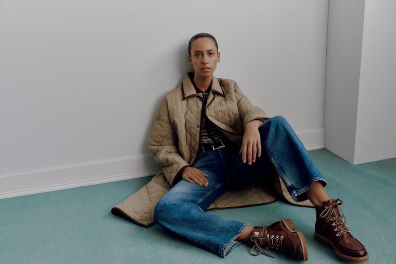 Model seated on pale teal carpet wearing a beige quilted coat, blue jeans, and brown leather boots against a white wall.