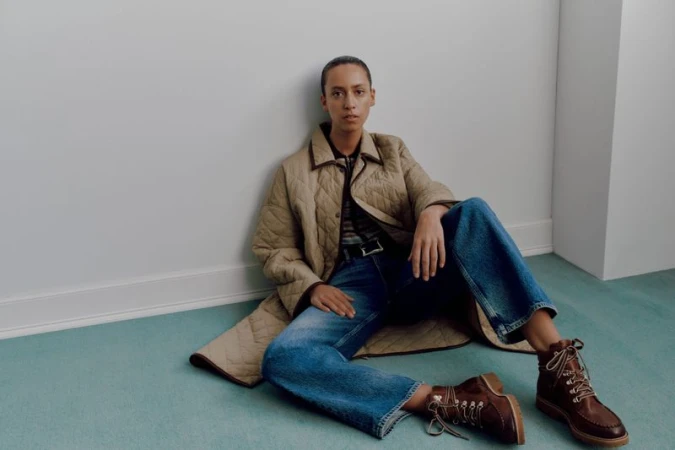 Model seated on pale teal carpet wearing a beige quilted coat, blue jeans, and brown leather boots against a white wall.