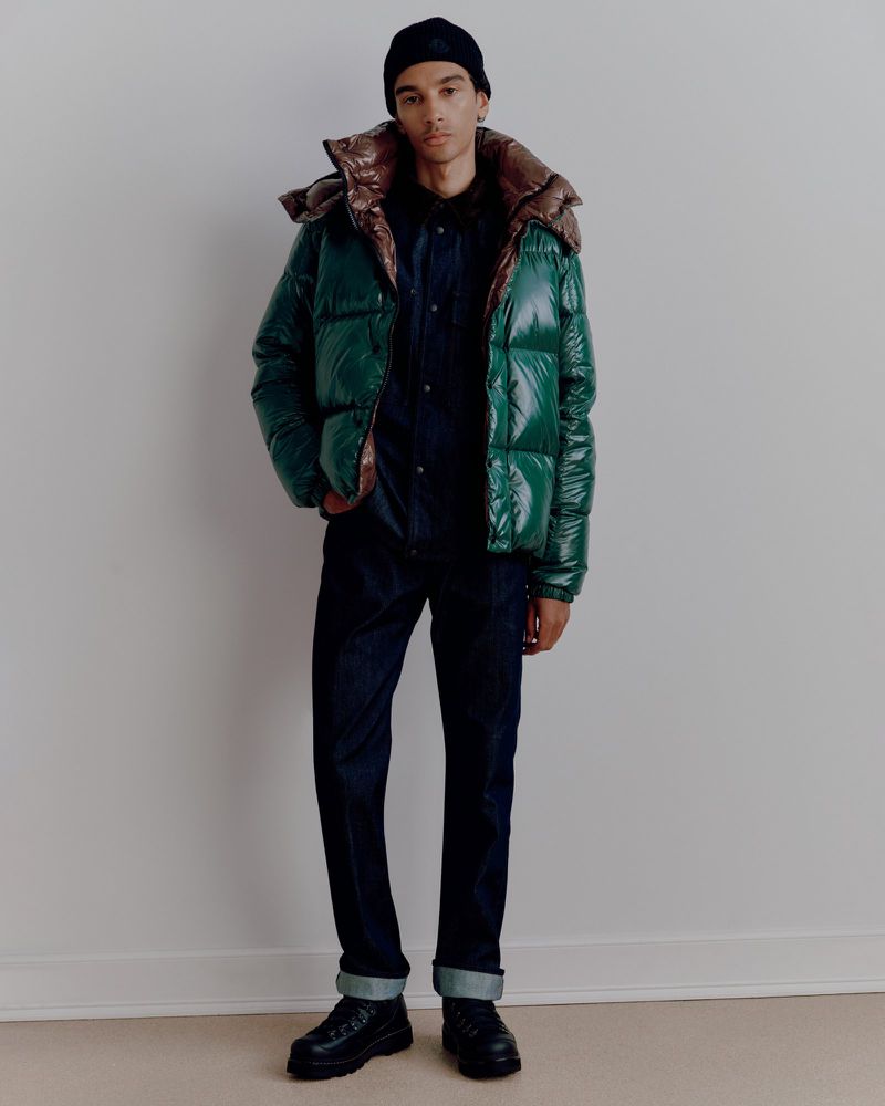 Full-length shot of an adult male in a green puffer jacket, black beanie, dark shirt and pants, and black boots against a plain wall.