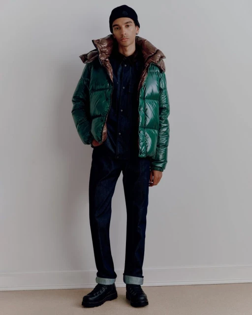 Full-length shot of an adult male in a green puffer jacket, black beanie, dark shirt and pants, and black boots against a plain wall.