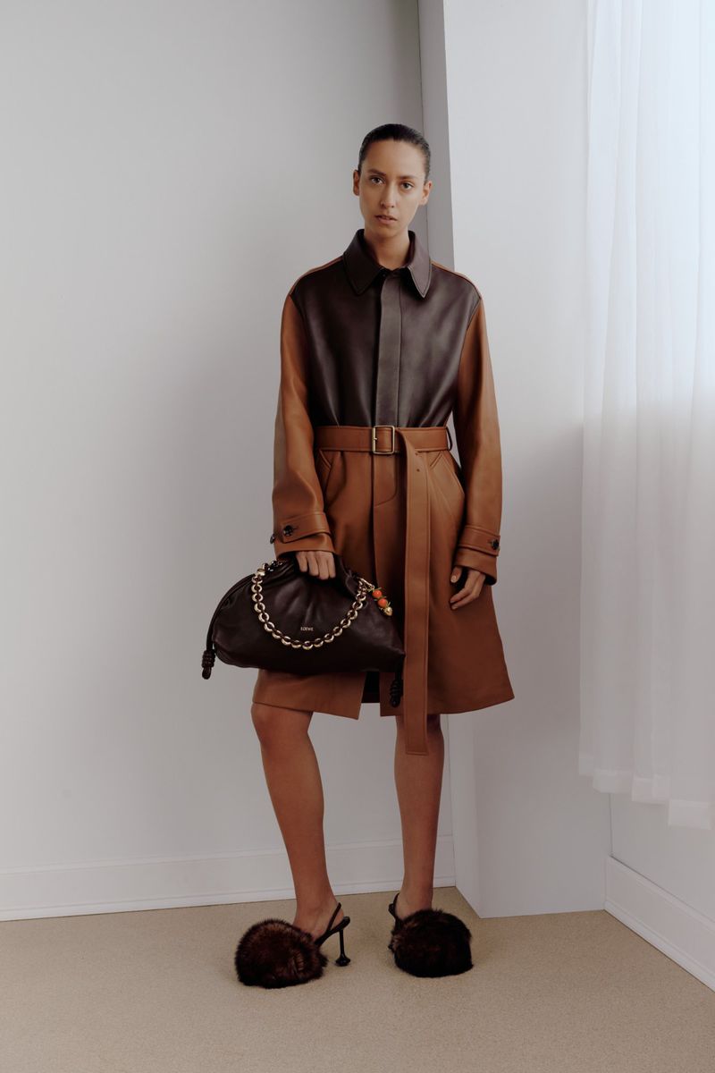 A woman poses in a brown two-tone coat, holding a dark handbag, with brown furry heeled shoes, against a plain corner studio backdrop.