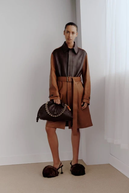 A woman poses in a brown two-tone coat, holding a dark handbag, with brown furry heeled shoes, against a plain corner studio backdrop.