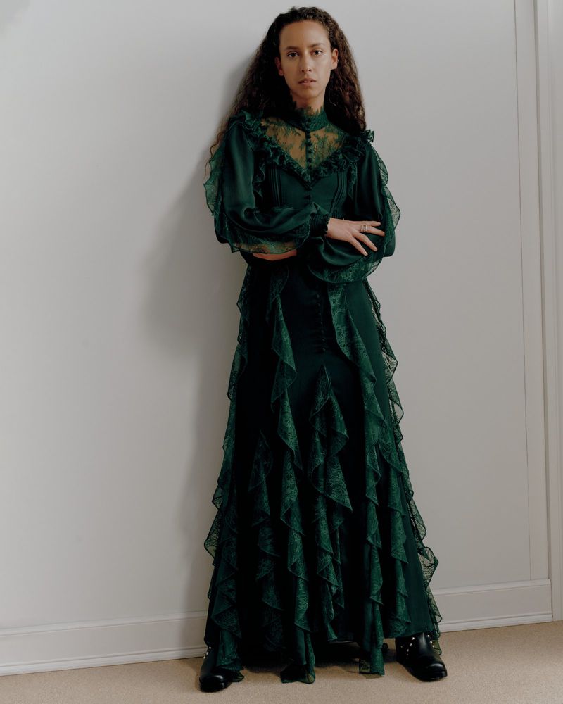 A woman with curly hair stands indoors, modelling a long emerald green dress with lace details, puffed sleeves and black shoes.