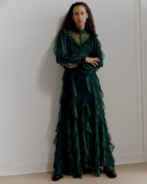 A woman with curly hair stands indoors, modelling a long emerald green dress with lace details, puffed sleeves and black shoes.