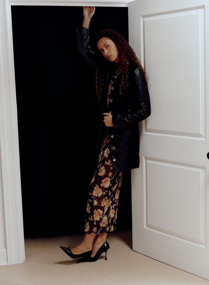 A woman poses in an open doorway, wearing a black jacket over a floral print dress and black high heel shoes, against a dark backdrop.