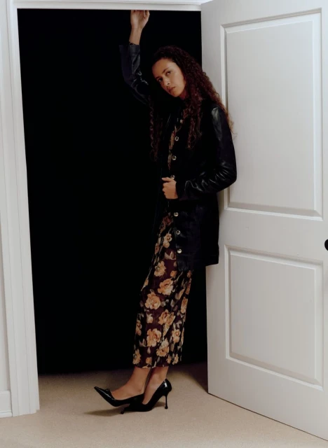 A woman poses in an open doorway, wearing a black jacket over a floral print dress and black high heel shoes, against a dark backdrop.