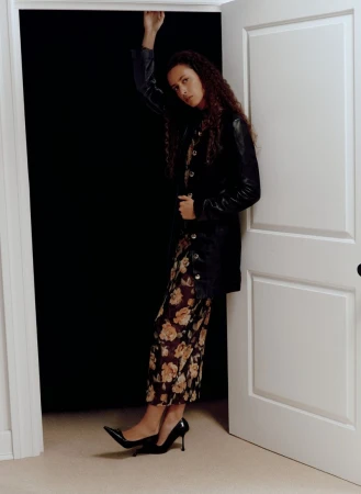 A woman poses in an open doorway, wearing a black jacket over a floral print dress and black high heel shoes, against a dark backdrop.