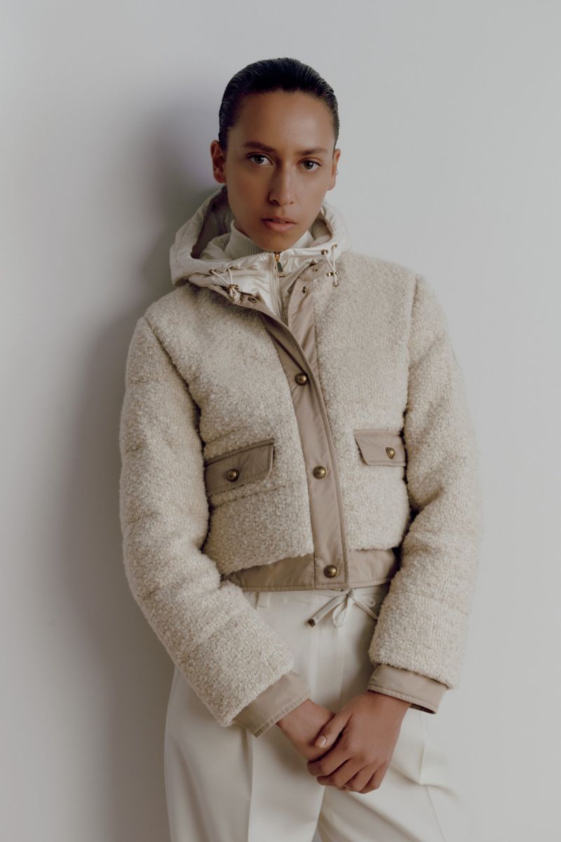 A woman wears a beige hooded teddy jacket and ecru-colored pants, posing against a plain backdrop in a fashion style studio shot.