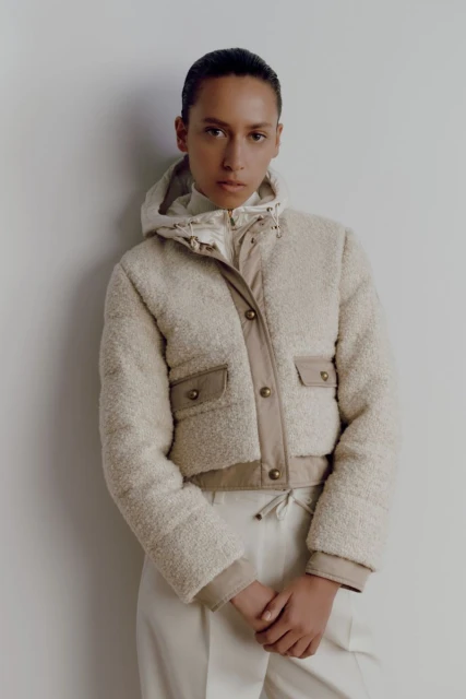 A woman wears a beige hooded teddy jacket and ecru-colored pants, posing against a plain backdrop in a fashion style studio shot.