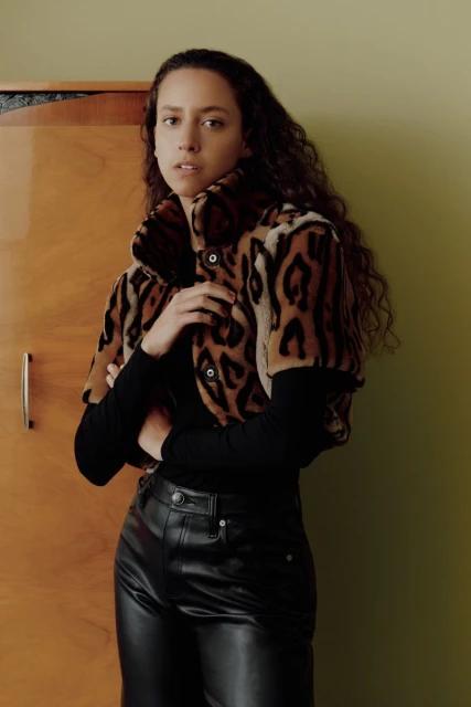 Brunette woman in a leopard-print jacket and black leather pants poses indoors against a neutral wall, looking at the camera.
