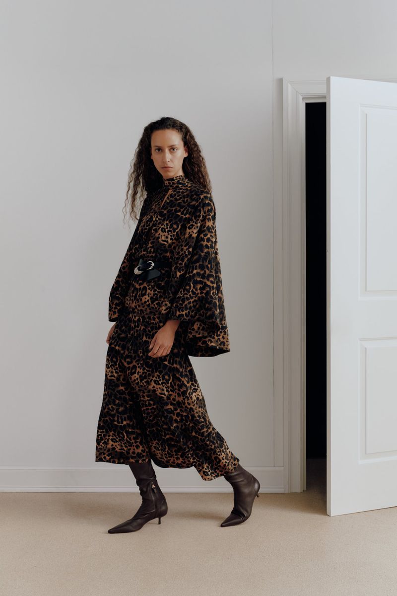Fashionable woman poses indoors wearing a leopard-print midi dress, wide black belt, and ankle boots in a fashion editorial shot.