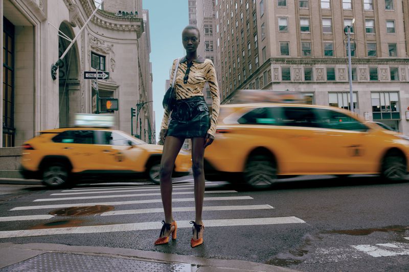Fashion shot of an adult female in patterned top and skirt standing on a crosswalk in a city street with yellow taxis in the background.