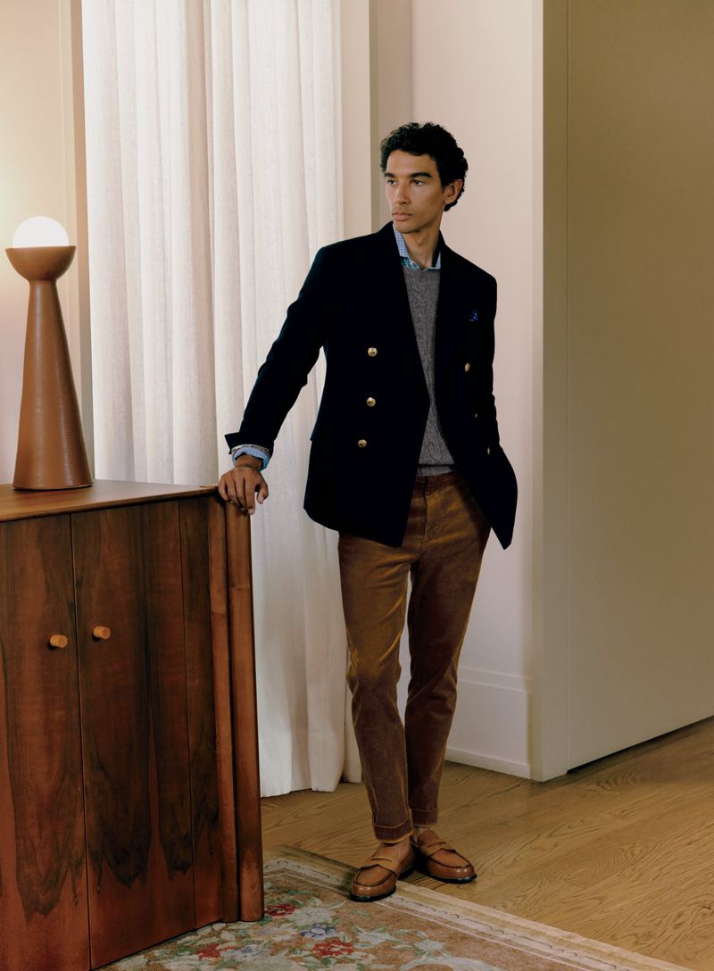 Man in a navy blazer and brown corduroy pants stands indoors, leaning against wooden cabinet with a lamp, gazing out of frame.