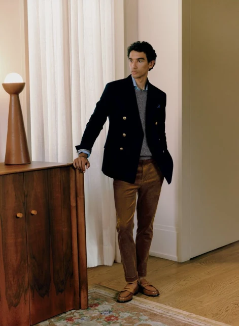Man in a navy blazer and brown corduroy pants stands indoors, leaning against wooden cabinet with a lamp, gazing out of frame.