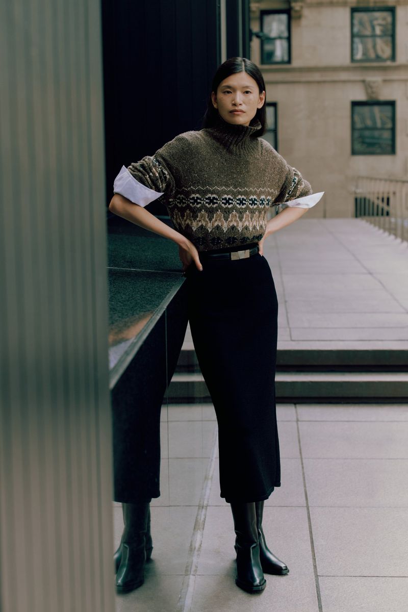 Asian woman poses in a brown patterned sweater, black skirt, and black boots on an outdoor patio with a city building background.