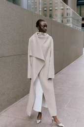 Model wearing a long cream coat and patterned heels posing outdoors near a large stone wall for Saks Fall Fashion