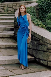 A red-haired model in a long draped cobalt blue dress standing on outdoor stone steps while holding a small patterned clutch bag.