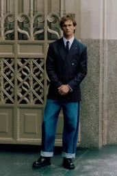 A young male model wearing a navy pinstripe blazer and cuffed denim jeans standing against an ornate metal gate.