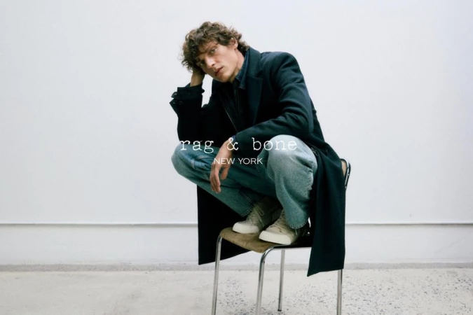 A young adult male with curly brown hair posing while seated on a chair, wearing jeans, sneakers, and a long coat in a photo studio.