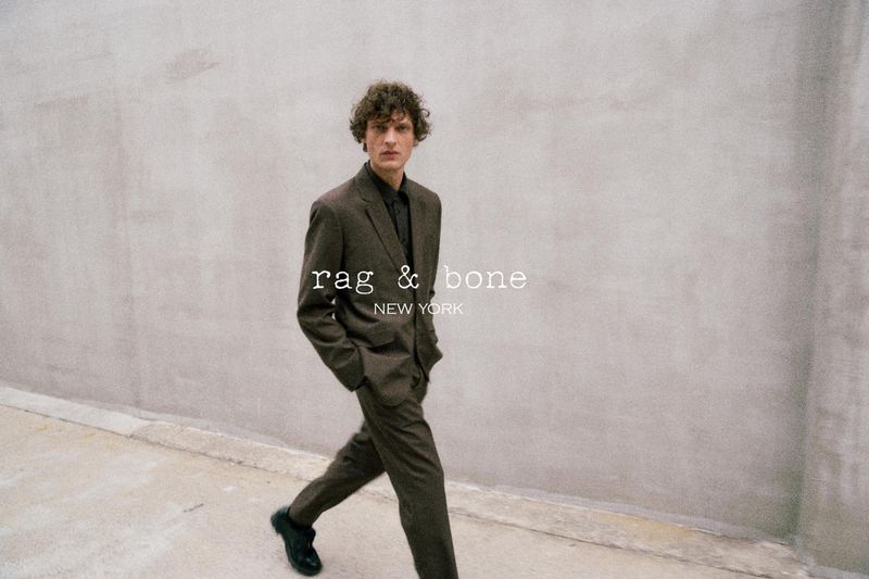 Man in brown suit walks along grey city sidewalk. Rag & Bone logo on wall. Fashion editorial shot with neutral tones and natural light.