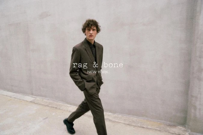 Man in brown suit walks along grey city sidewalk. Rag & Bone logo on wall. Fashion editorial shot with neutral tones and natural light.