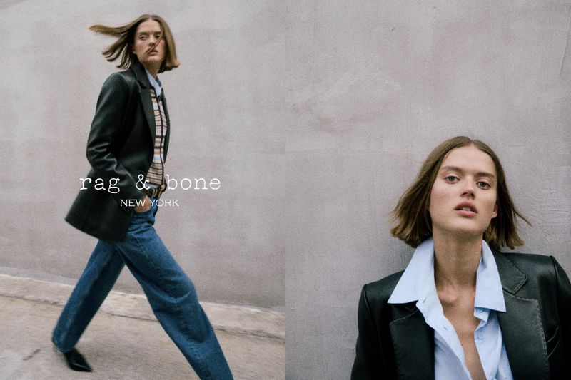 Fashion advertisement featuring a female model wearing a blazer, shirt, and jeans, posing in front of a grey wall, brand logo visible.