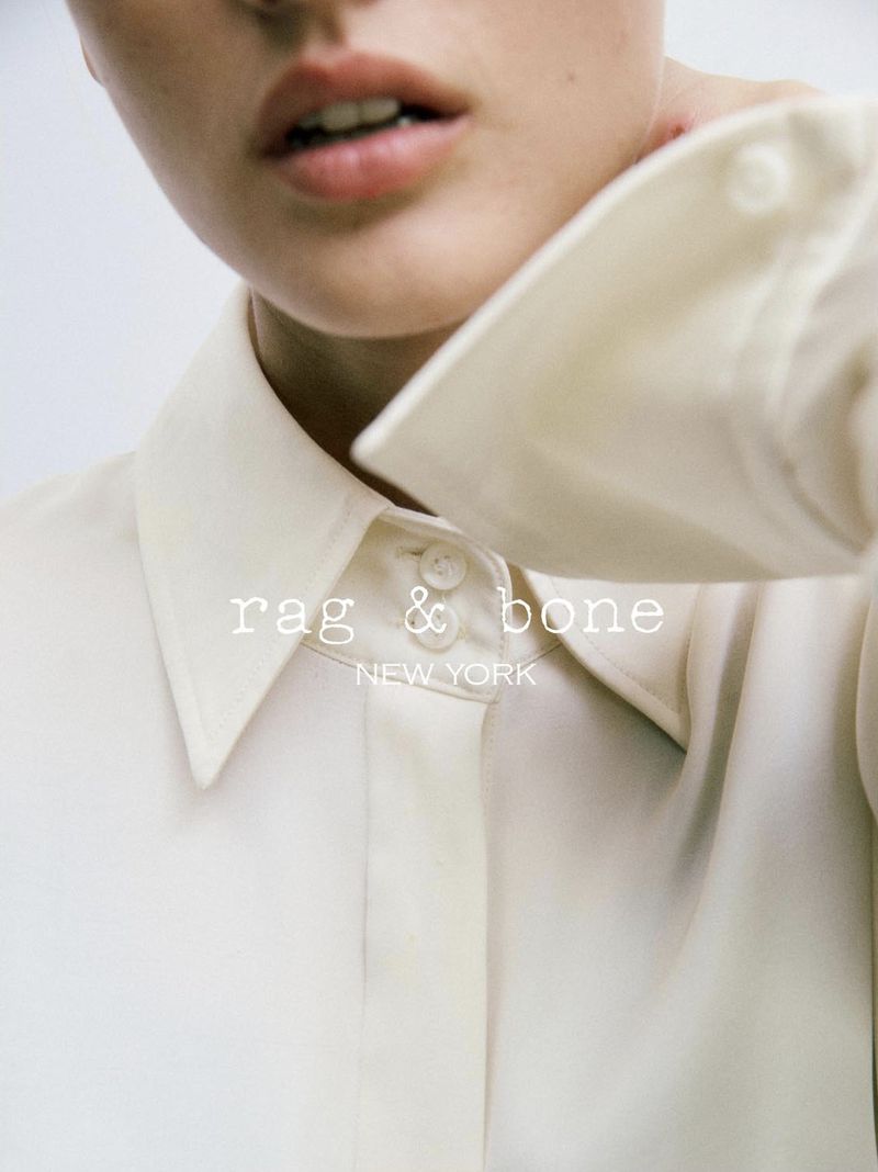 Close-up of an adult female wearing a white, collared shirt with "rag & bone NEW YORK" logo overlaid on the garment in white font.
