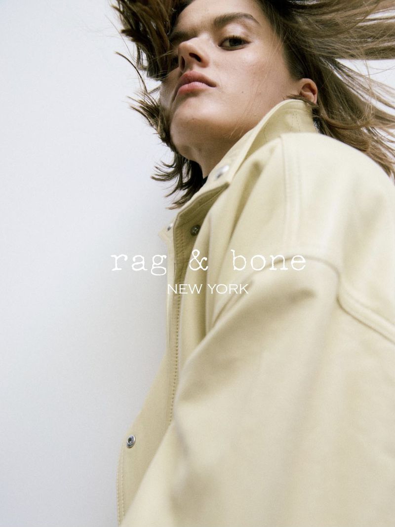 Fashion portrait of an adult female model wearing a jacket looking at the camera with flowing hair, against a white background.