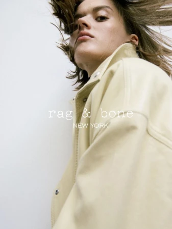 Fashion portrait of an adult female model wearing a jacket looking at the camera with flowing hair, against a white background.
