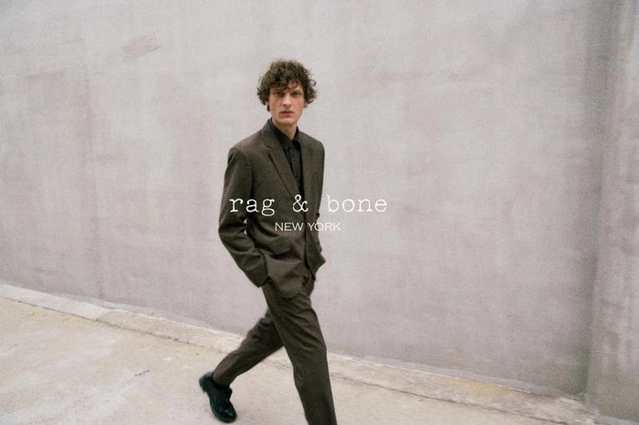 Model Valentin Caron walking in a dark tailored suit along a neutral concrete wall for the Rag & Bone FW 25 campaign.