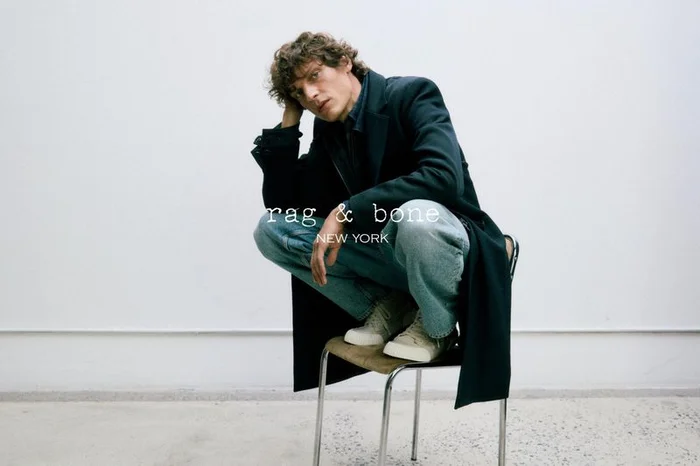 Male model crouched on a chrome chair wearing a dark coat and blue jeans against a white background