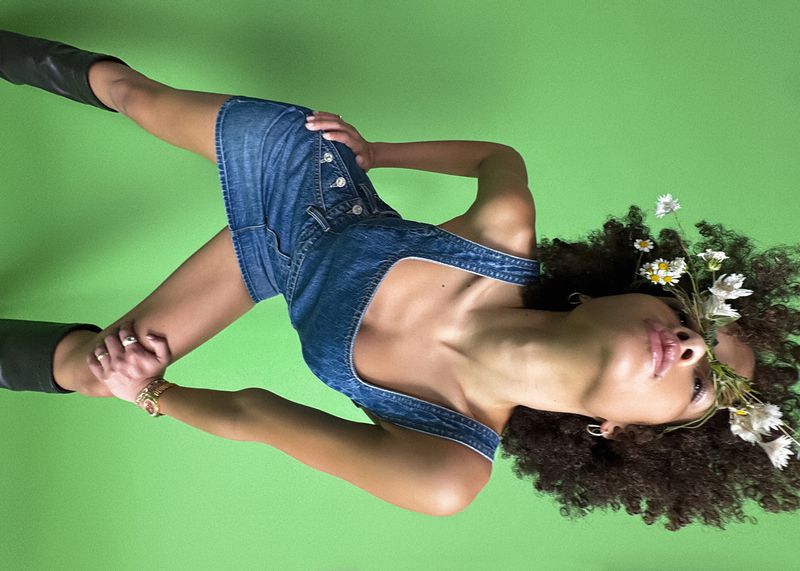 An adult female poses in a denim dress and black boots, adorned with flowers, against a green background, showcasing a fusion of fashion and nature.