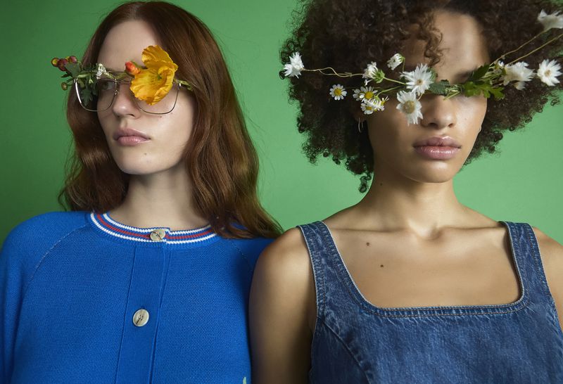 Two women wearing floral eyeglasses against a green backdrop. One woman wears a blue sweater, and the other is in a denim top.