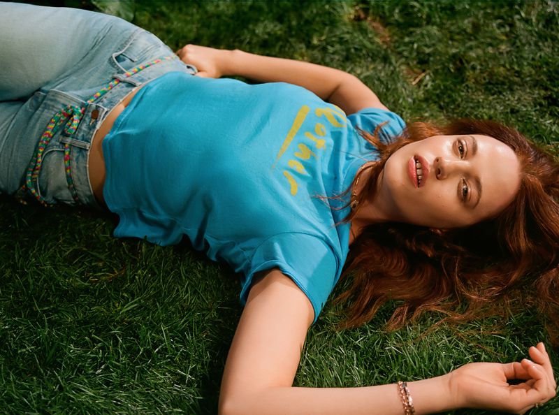 Young woman with auburn hair lies on green grass, wearing a blue T-shirt and blue jeans in an outdoor setting with natural lighting.