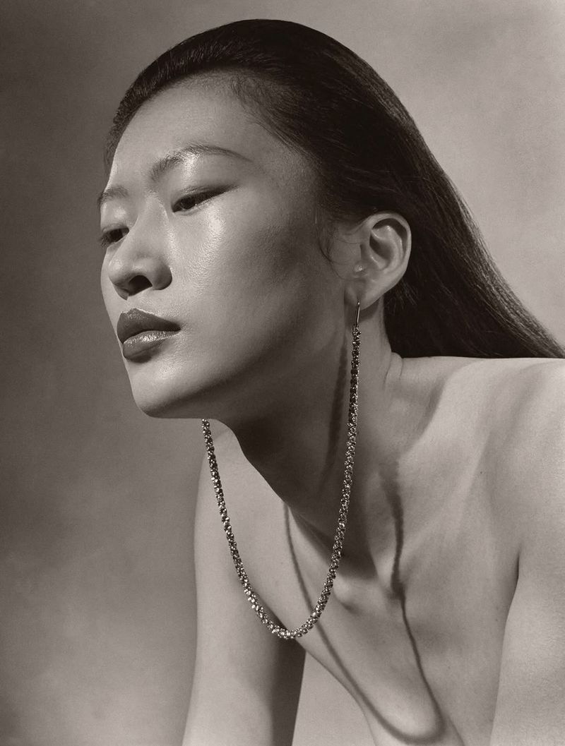 Asian woman with bare shoulders poses in a photo studio with long dangling earrings. The image has a clean background and neutral tones.