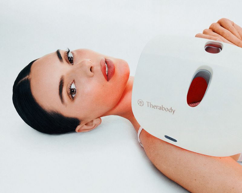Attractive woman with dark hair poses with a white Therabody device against a bright white backdrop in a beauty and health setting.