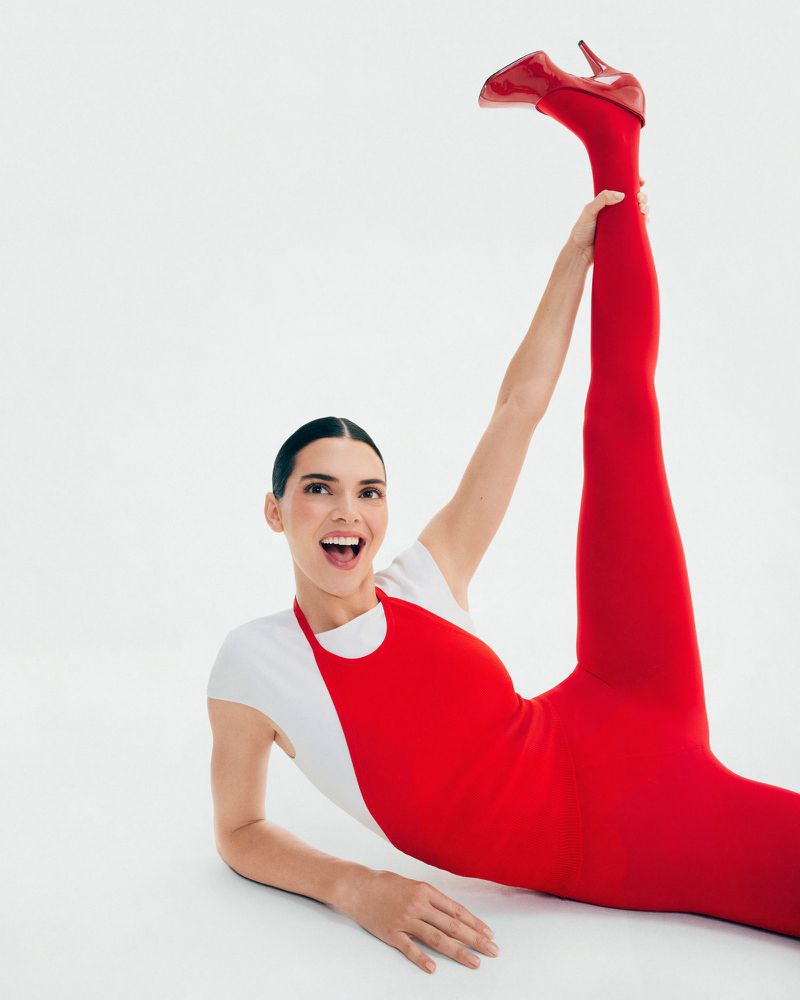 A woman lies on a white background wearing a white shirt, a red leotard, red leggings, and red high heels with one leg raised.