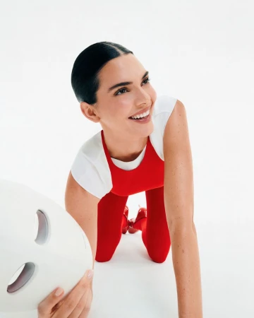 A woman with dark hair smiles while kneeling on a white surface next to a light therapy mask, facing toward the upper right.