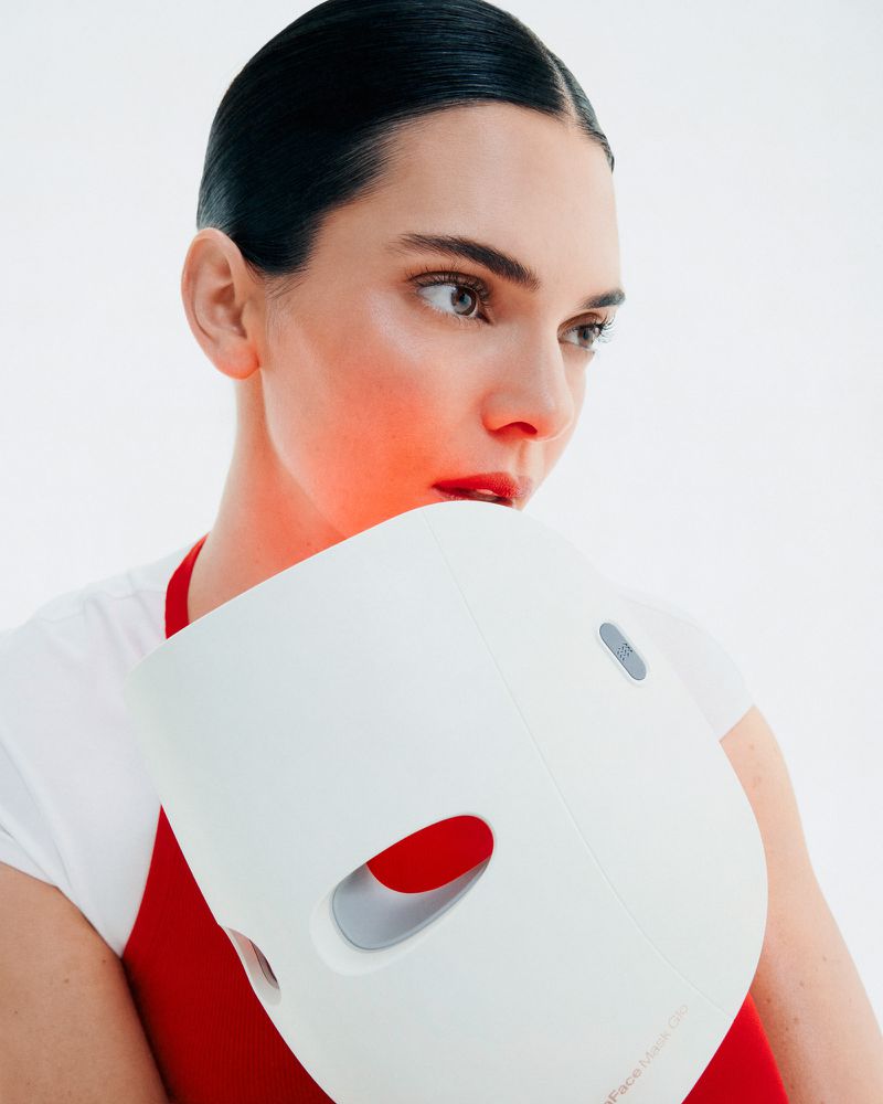 Close-up of a young woman with red lipstick holding a white LED face mask, looking away in a minimalist white studio setting.