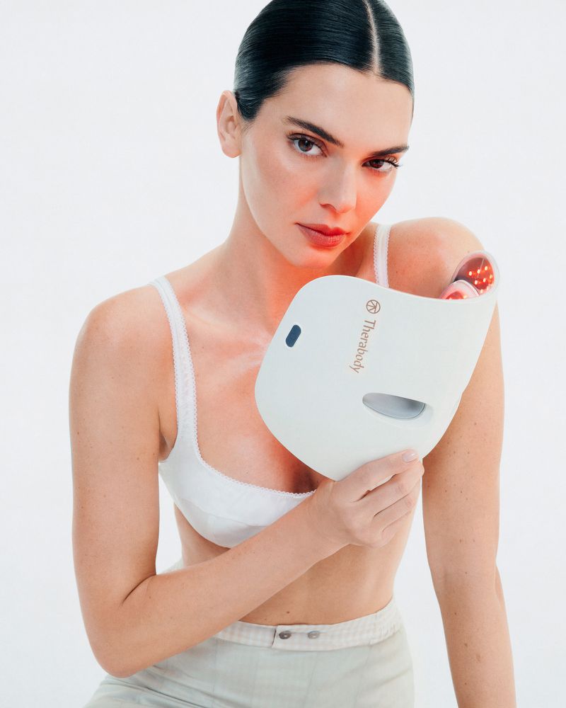 Attractive woman poses with a Therabody LED face mask. She wears a bra top and low-rise pants against a plain white background.