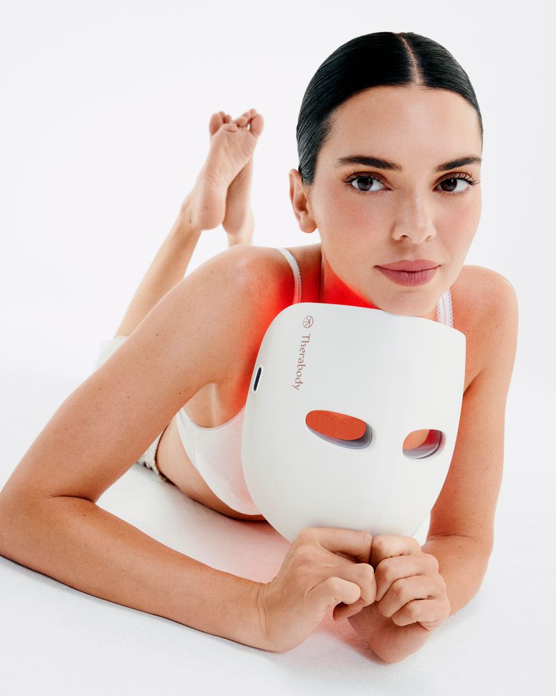 Adult female lies on white surface holding a white light therapy mask, illuminated in red, with dark hair slicked back.