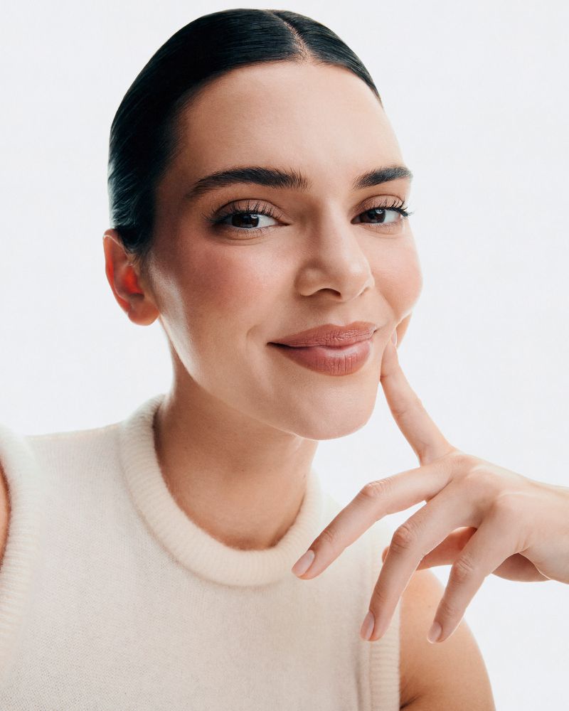Close-up portrait of an adult woman with dark hair pulled back, wearing a cream-colored top, touching her cheek with her finger, smiling softly.