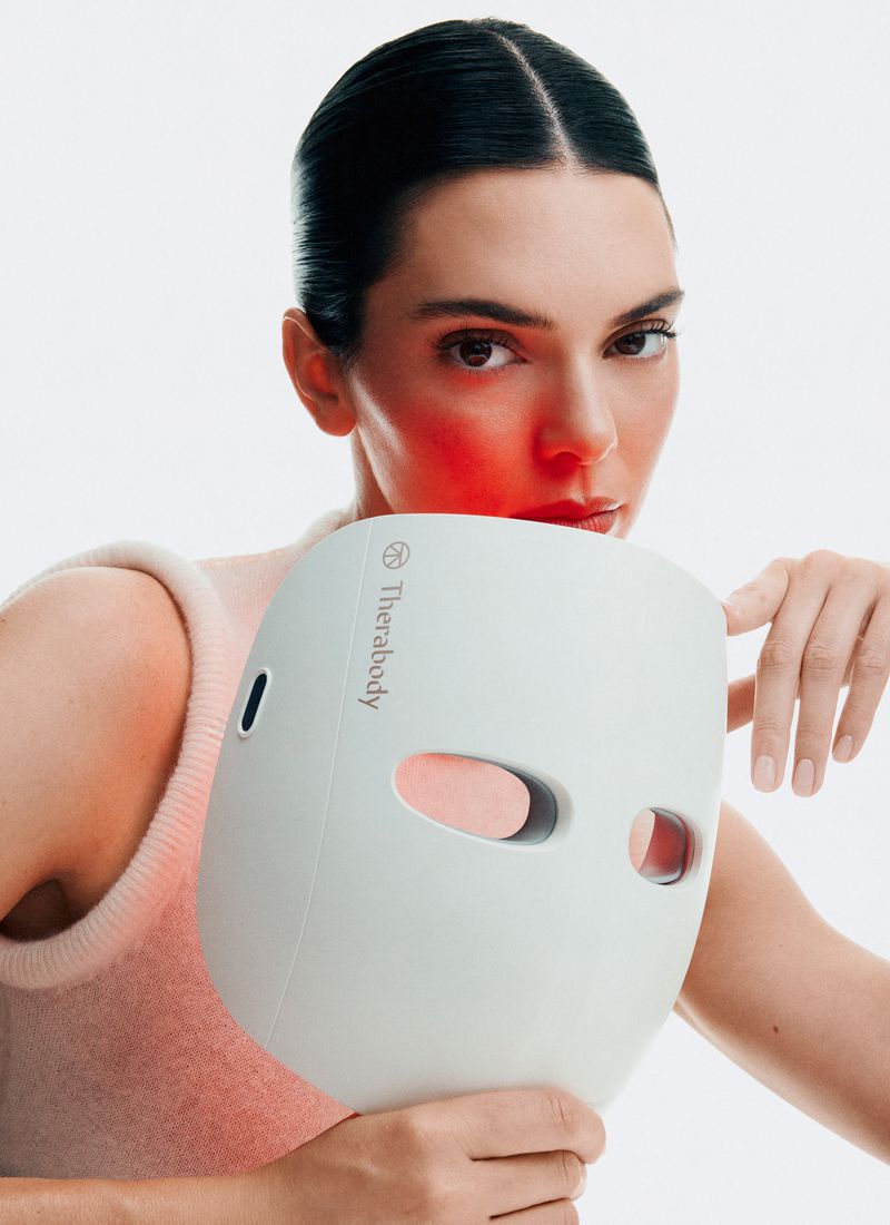 Woman holds Therabody LED face mask, fair skin, dark hair, wearing a light top against a white background, red LED light glow.