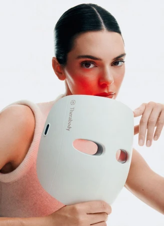 Woman holds Therabody LED face mask, fair skin, dark hair, wearing a light top against a white background, red LED light glow.