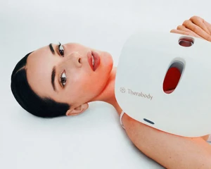 Kendall Jenner lying down against a white background holding a white Therabody device over her shoulder.