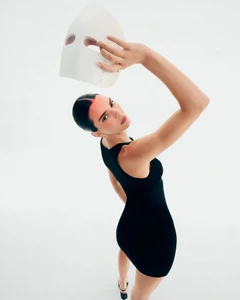 Kendall Jenner in a black sleeveless mini dress holding a white LED face mask above her head on a white backdrop.