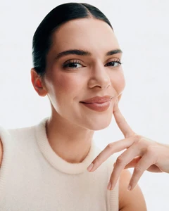 Kendall Jenner smiling and resting fingers on her cheek wearing a cream knit top against a white studio background.