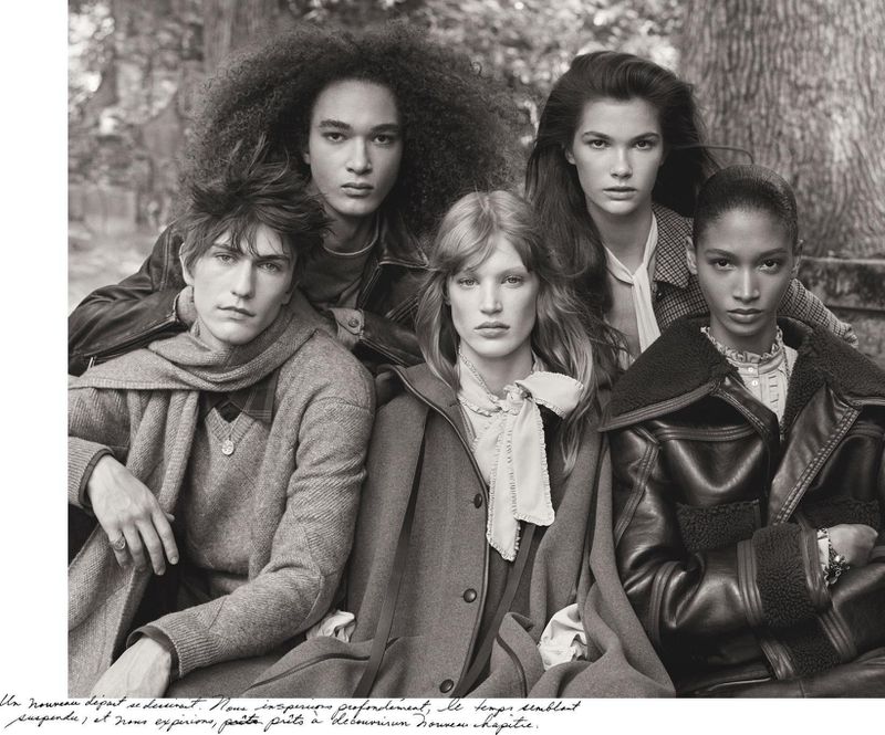 Black and white image of a group of five young adults sitting close together outside, wearing coats and sweaters in a sombre mood.
